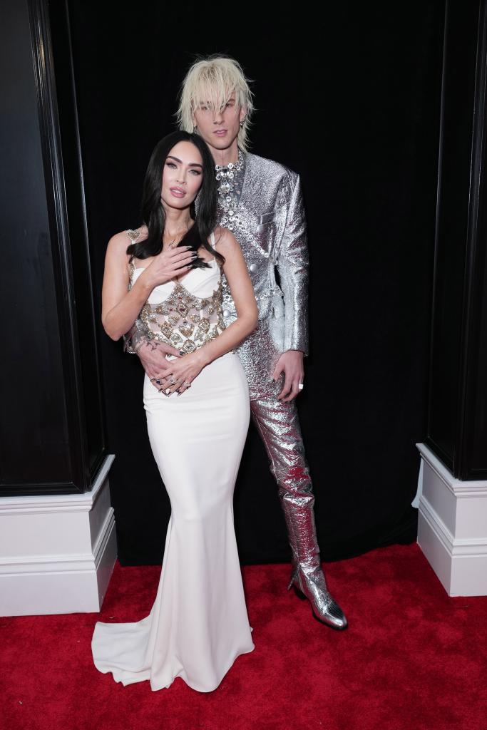 Machine Gun Kelly wearing a silver suit while putting his arm around Megan Fox in a white dress on a red carpet.