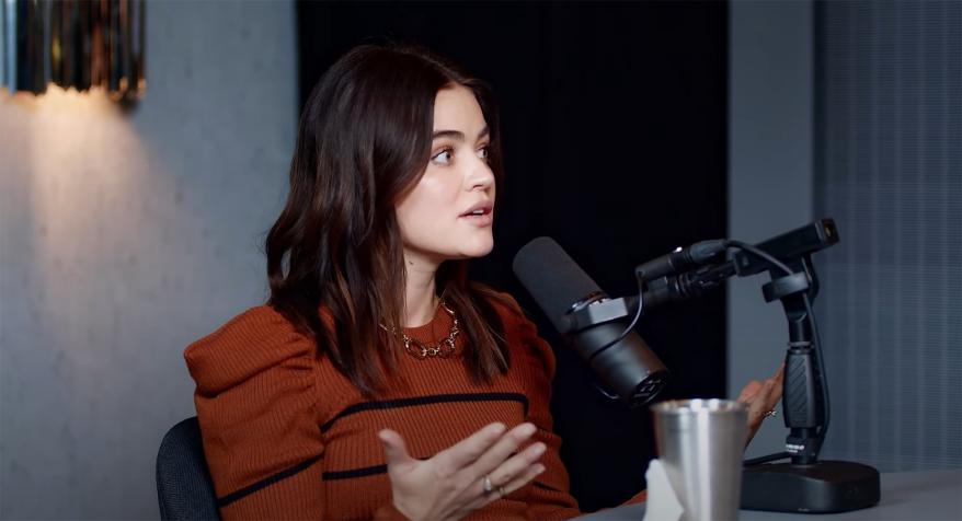 Lucy Hale speaks into microphone in red sweater and chain necklace