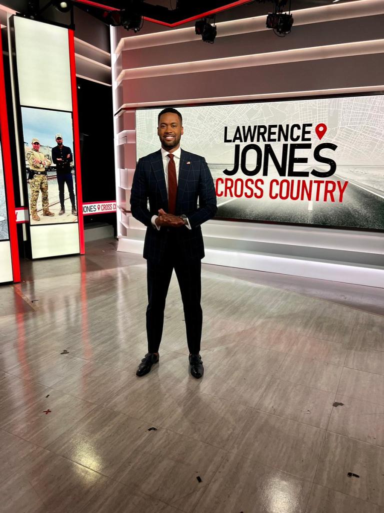 Fox News anchor, wearing a dark suit and red tie, stands on the set of his cable show "Lawrence Jones Cross Country."