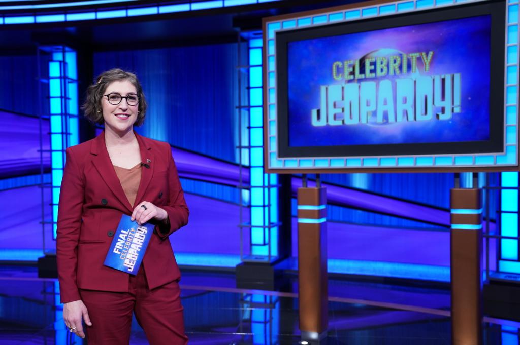 mayim bialik posing next to a "celebrity jeopardy" sign