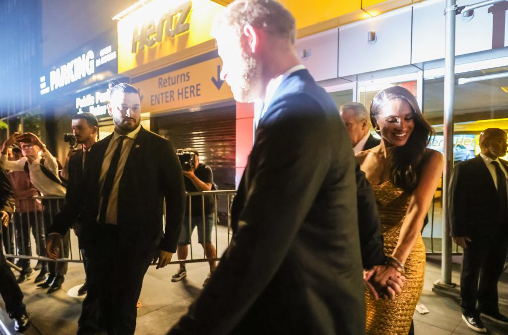 Prince Harry, wearing a dark suit, and Meghan Markle, in a gold strapless dress, are flashed by paparazzi photographers as they leave NYC's 19th precinct on Tuesday night