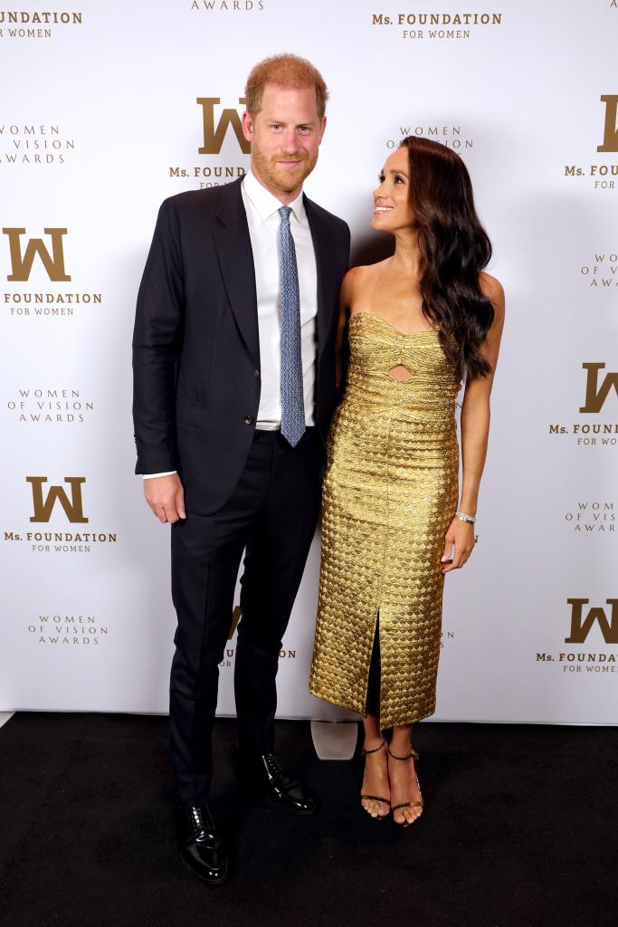 Prince Harry, wearing a dark suit and blue tie, poses on the red carpet with wife Meghan Markle, who is wearing a strapless gold dress