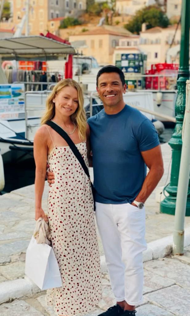 Kelly Ripa and Mark Consuelos stand in front of boats. Kelly in a printed maxi dress and Mark in white pants and a blue t-shirt.