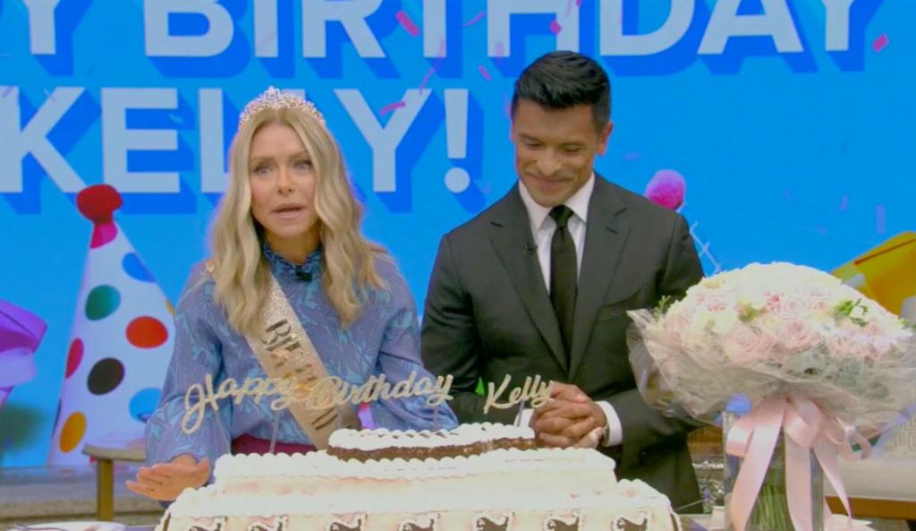 Kelly Ripa in a birthday crown and sash sits next to Mark Consuelos with a whale-shaped cake in front of her.