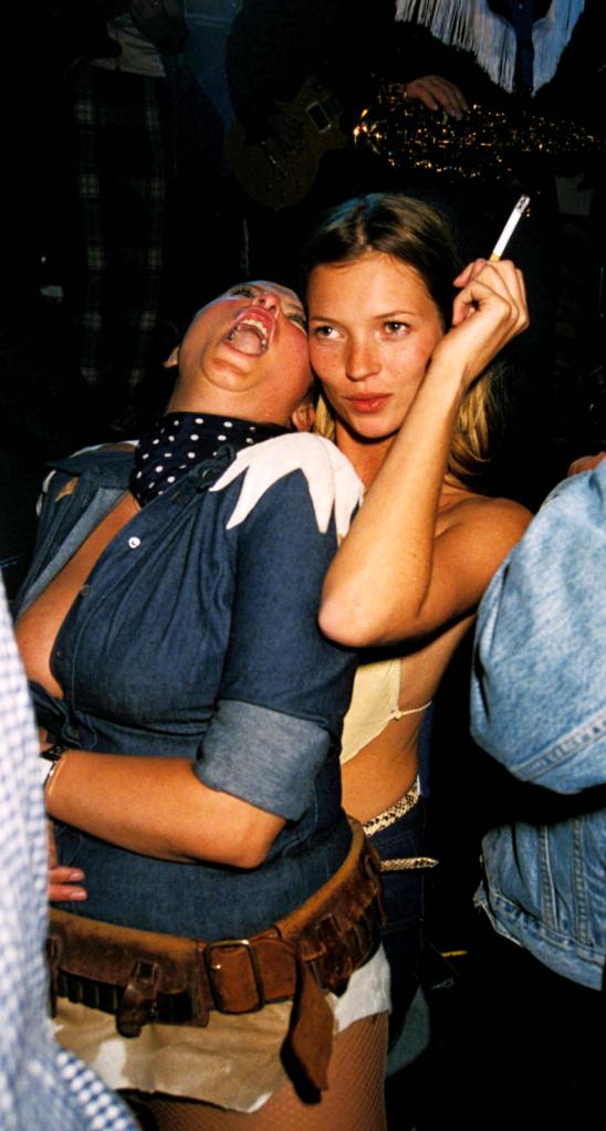 Kate Moss holding a cigarette while a pal leans on her.