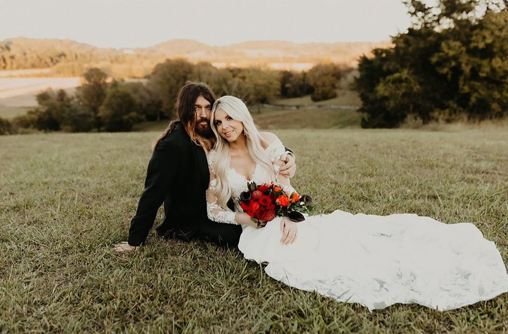 Billy Ray Cyrus and Firerose, in wedding attire, sitting in a field.