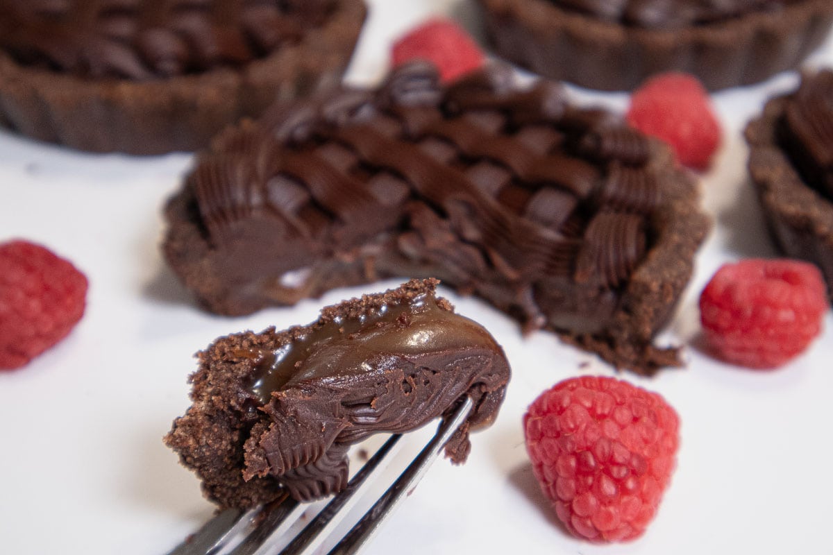 This shot of the Chocolate Caramel Truffle Tarts shows a forkful of the tart with a cut tart in the back ground with a few red raspberries.