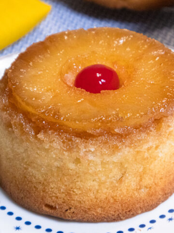 This Mini Pineapple Upside Down Cake features a pineapple slice with a cherry in the middle on top of a yellow cake sitting on a blue and white plate.