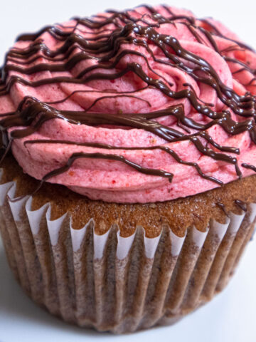 A lone Banana Split Cupcake with its vibrant pink strawberry buttercream and drizzled with chocolate sits on a pink rimmed plate.