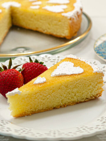 A slice of the Orange Almond Tea Cake sits on a white plate with 2 strawberries and the rest of the cut cake is behind it.