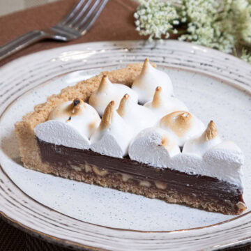 A slice of the S'more Tart with it's browned marshmallow topping sits on a brown rimmed plate with a fork and napkin in the background.