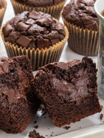 A cut open Double Chocolate Truffled Muffin sits on a plate with a teacup next to it and more muffins in the background.