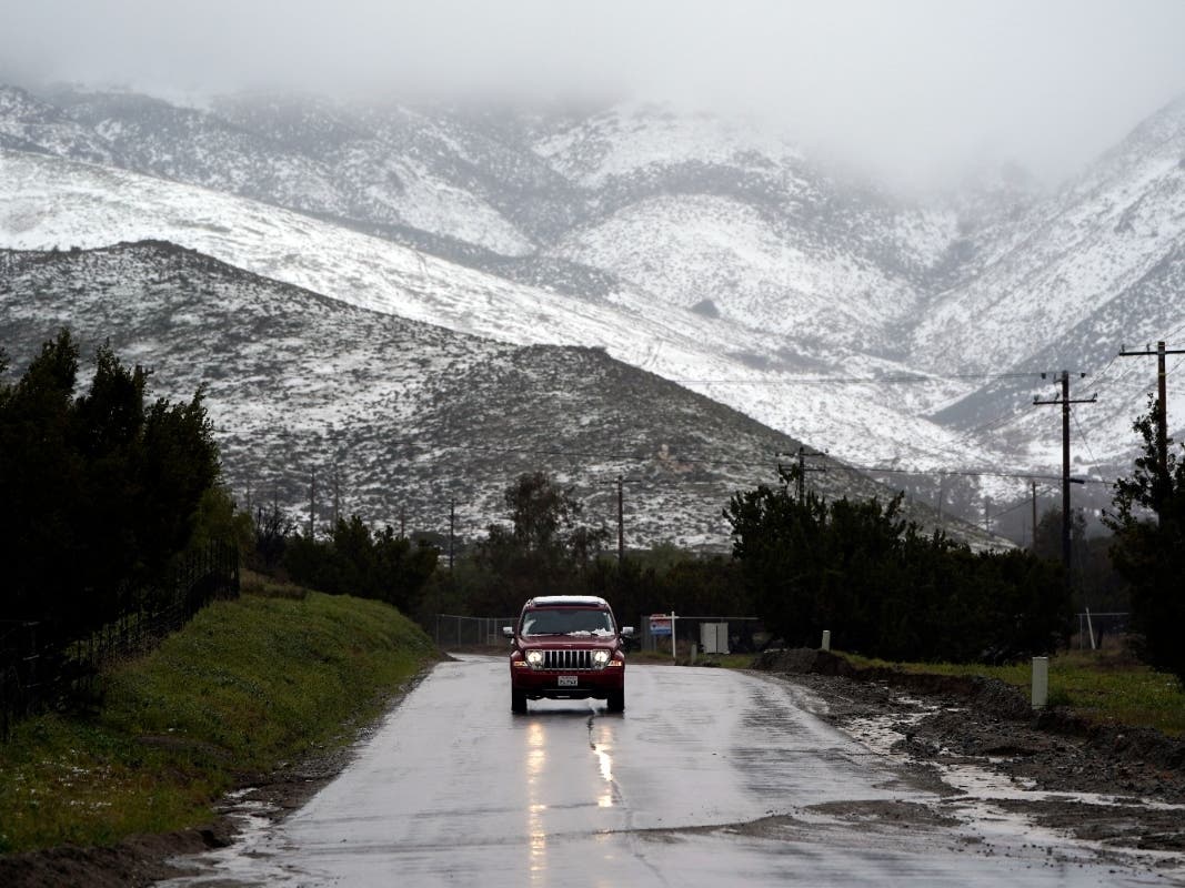 A motorist drives on a wet road under a snow-covered hillside Friday, Feb. 24, 2023, in Agua Dulce, Calif. California and other parts of the West faced heavy snow and rain Friday from the latest winter storm to pound the U.S. 