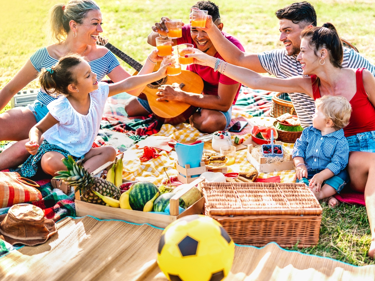 A family of children and adults toasting while enjoying an outdoor picnic. 