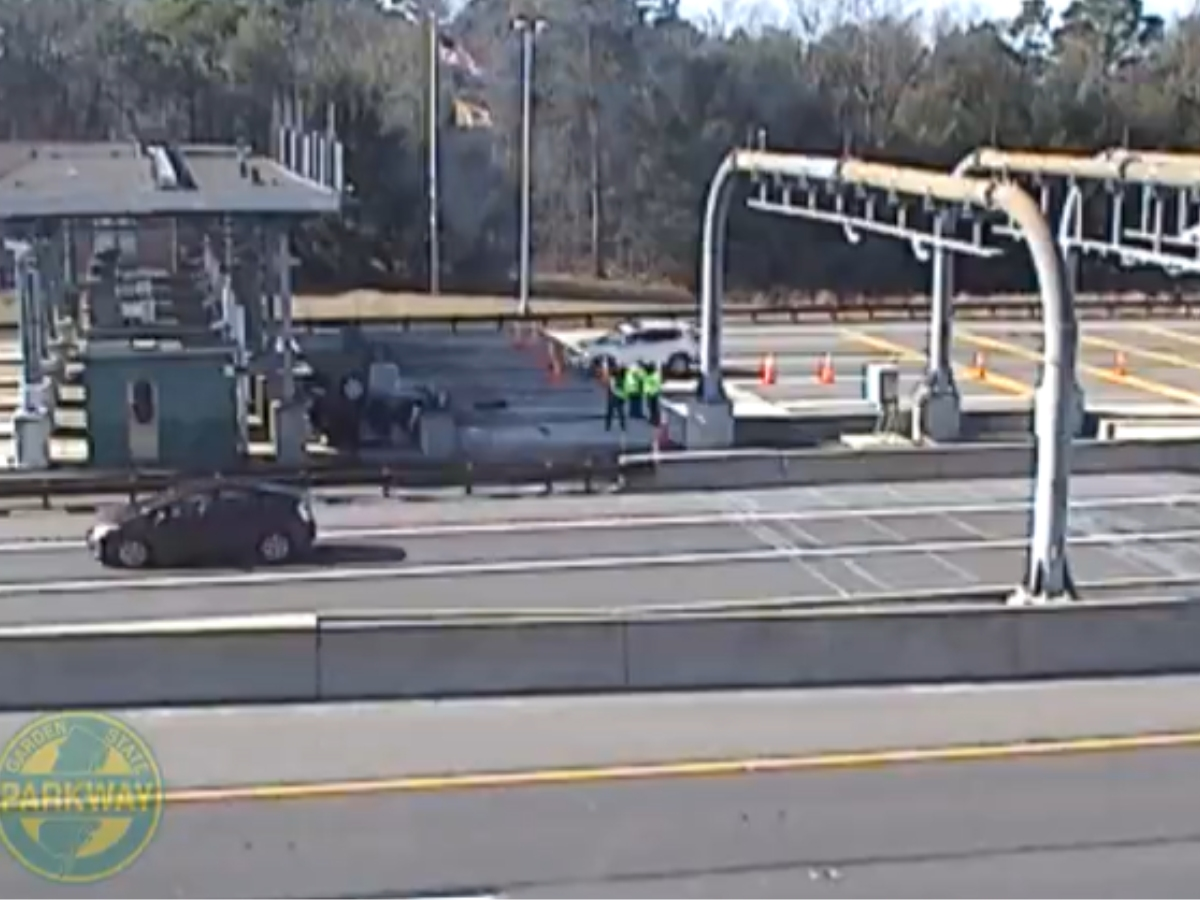 The toll booth camera shows the overturned car in the leftmost lane, with workers around it.