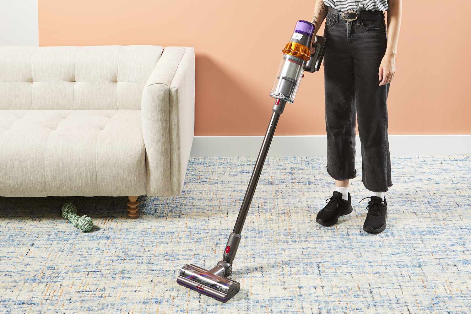 A person using a Dyson V15 Detect Cordless Vacuum Cleaner to clean a rug next to a white couch