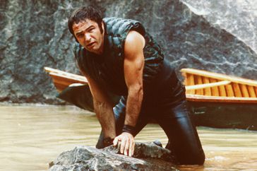 Burt Reynolds, US actor, holding onto some rocks as he struggles in the water with his boat behind him in a publicity still issued for the film, 'Deliverance'