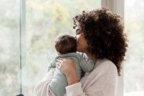 Young mom gives her baby a kiss on his head while standing next to a window in their home