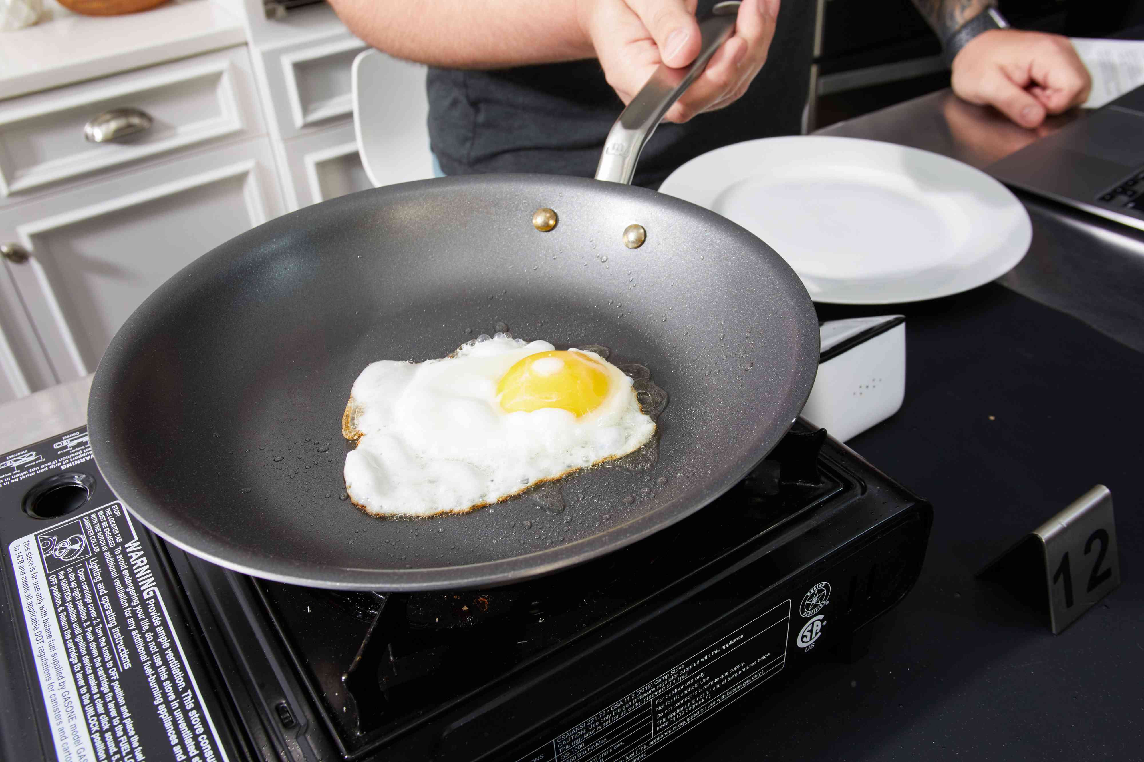 Person's hand holding handle of Made In nonstick sauté pan with egg cooking in oil