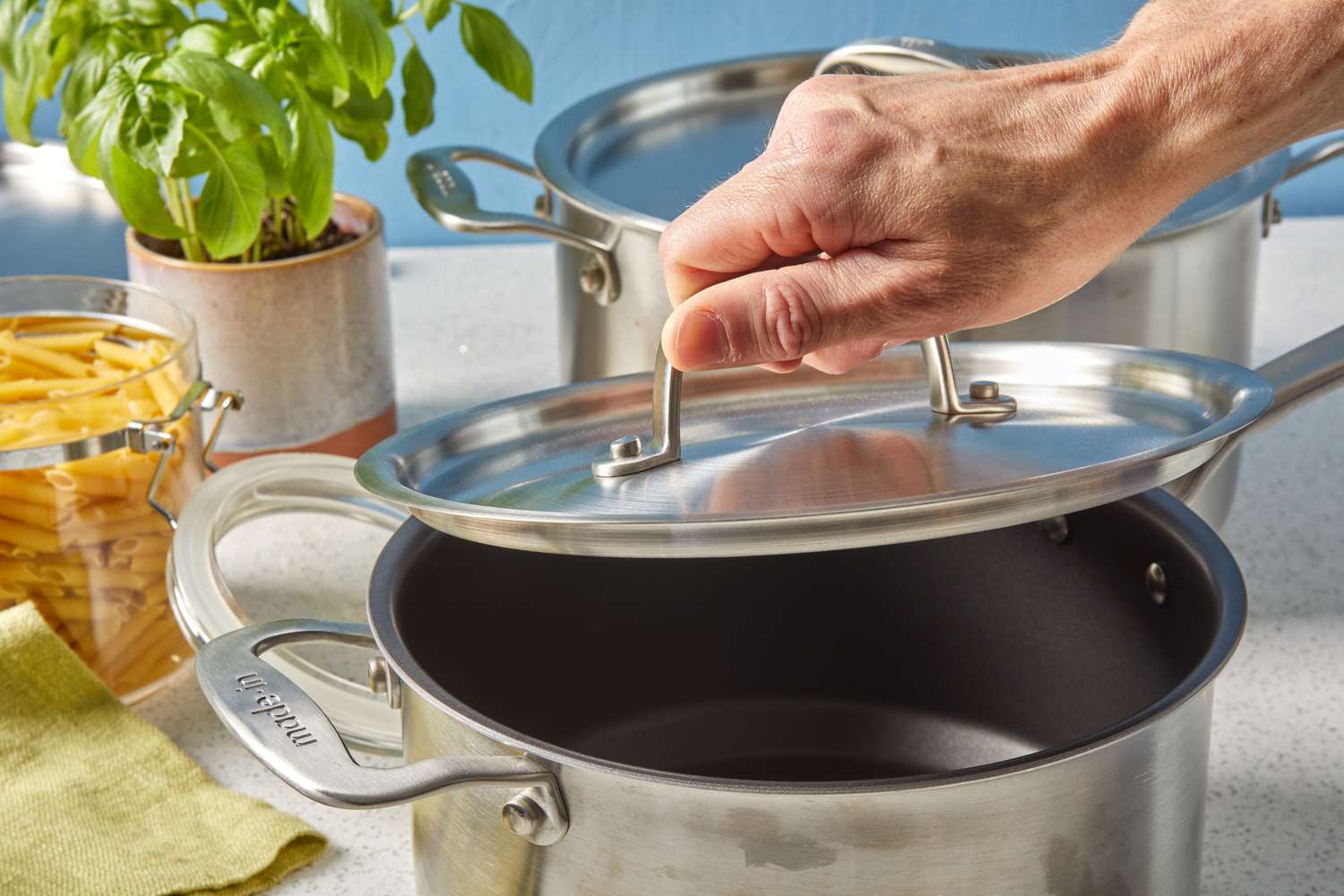 Hand lifting lid of Made In nonstick saucepan flanked by a basil plant and container of dry pasta 