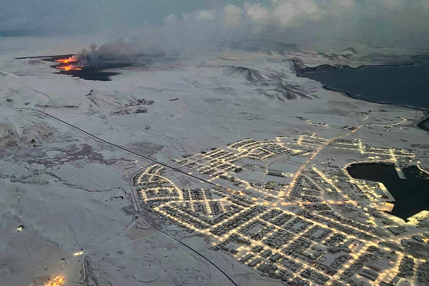 The evacuated Icelandic town of Grindavik (R) is seen as smoke billow and lava is thrown into the air from a fissure during a volcanic eruption on the Reykjanes peninsula 3 km north of Grindavik, western Iceland on December 19, 2023. A volcanic eruption began on Monday night in Iceland, south of the capital Reykjavik, following an earthquake swarm, Iceland's Meteorological Office reported. 