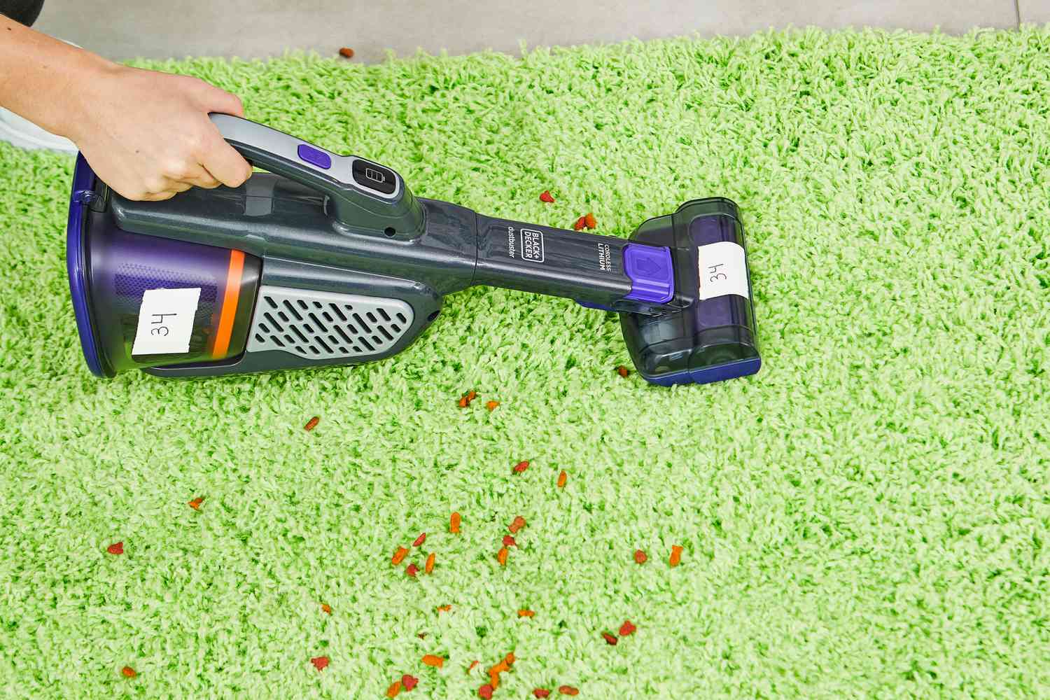 Hand using the Black+Decker Dustbuster AdvancedClean+ Pet Cordless Hand Vacuum Cleaner to clean pet food from a green carpet