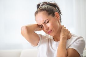 Woman with pain in the nech holding it with both arms with painful expression on her face.