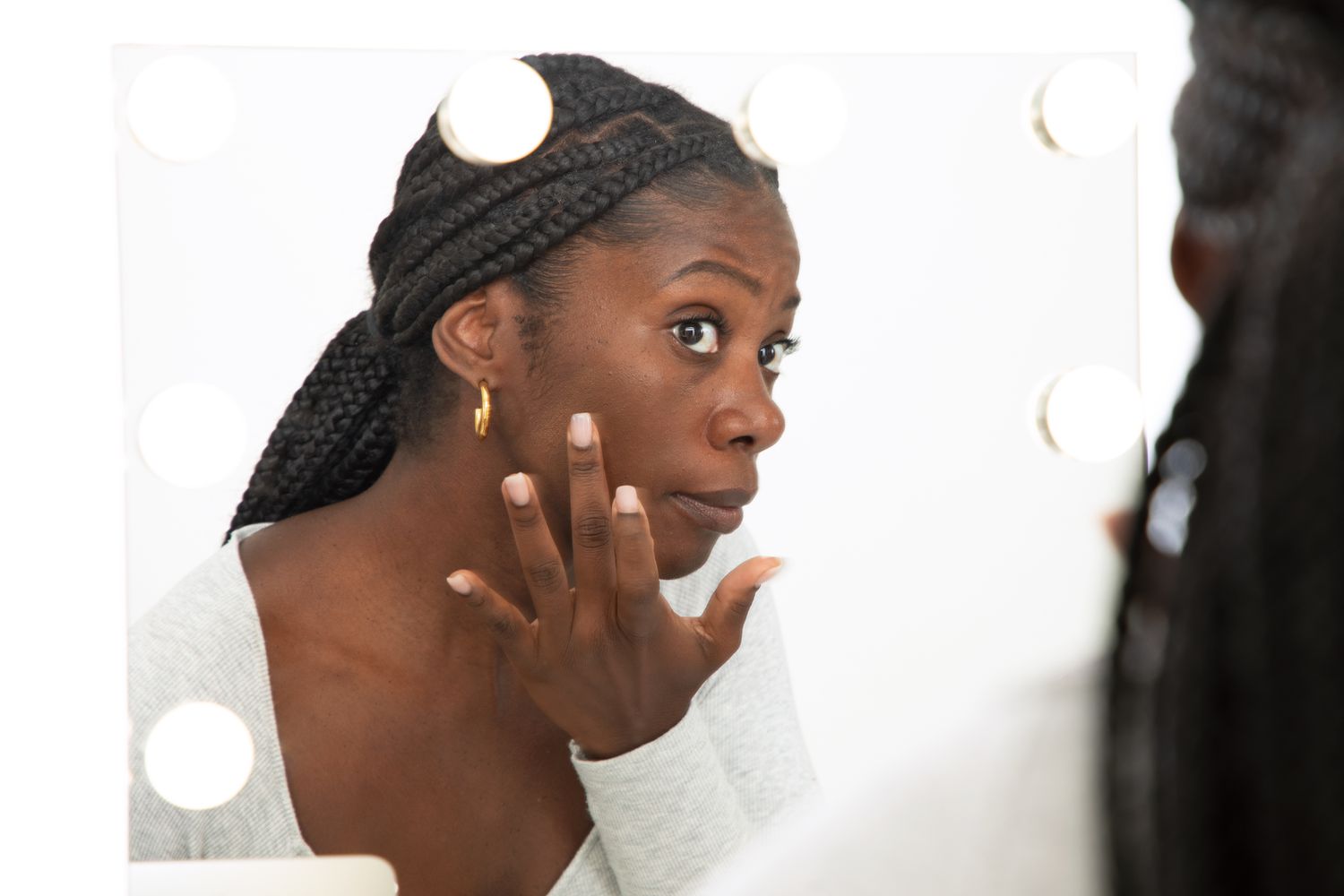 Woman applying HUDA BEAUTY GloWish Multidew Vegan Skin Tint Foundation to her face while looking into mirror