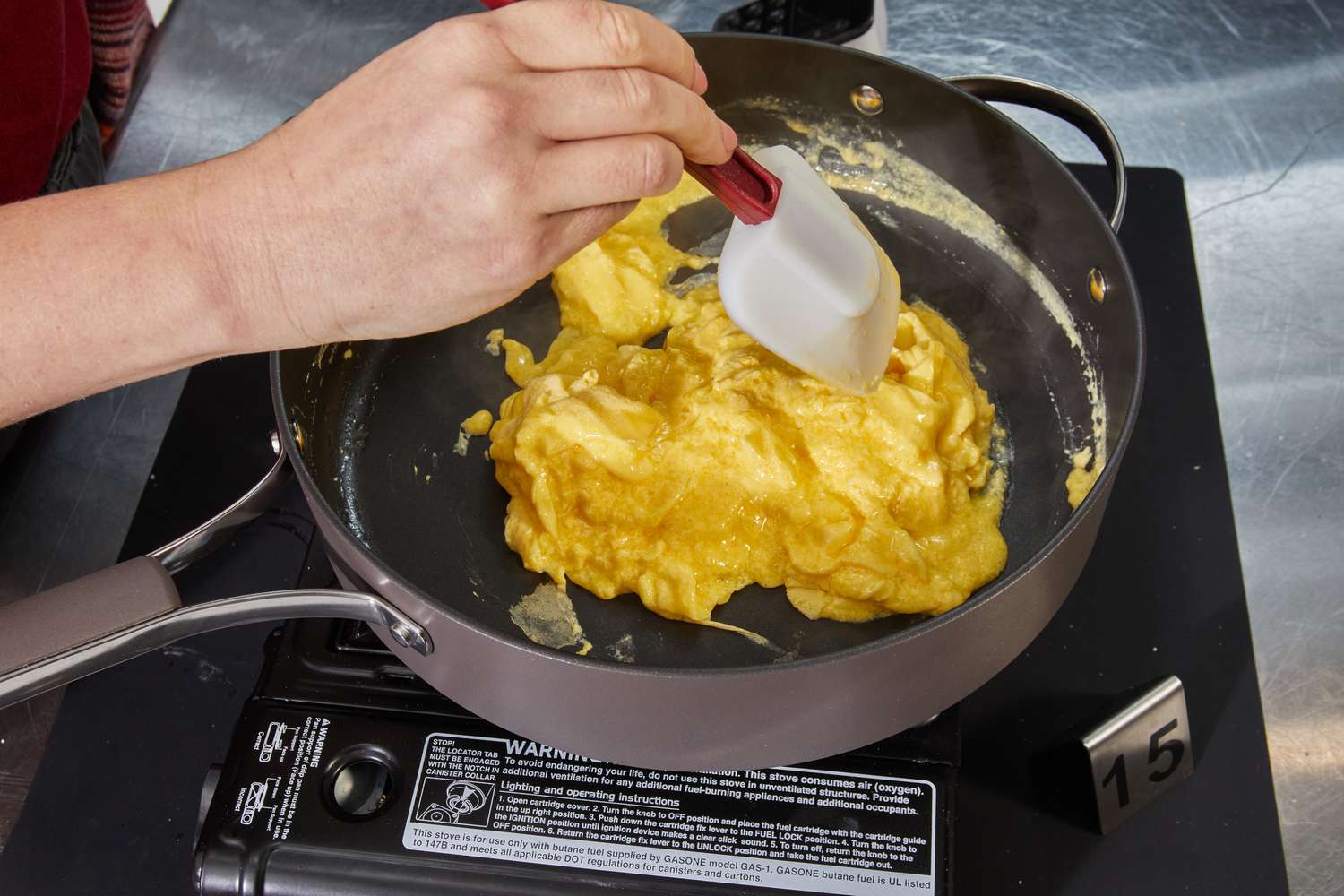 Hand using spatula to cook scrambled eggs in Ayesha Curry Hard-Anodized Nonstick 10-Piece Cookware Set saute pan