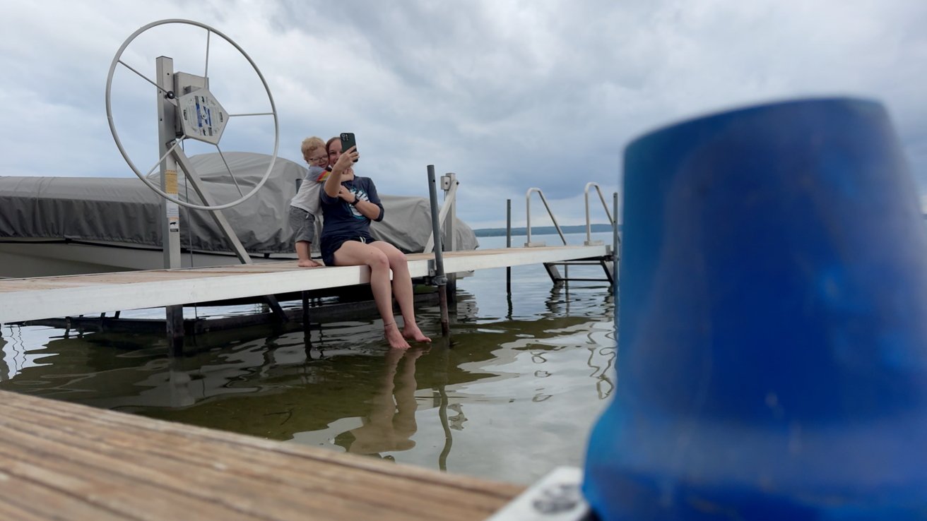 A photo of a woman and child sitting on a dock on a lake in front of a covered speed boat. They're taking a selfie together with a buoy out of focus in the foreground.