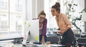 Two woman leaning over their desk looking at their computer monitor