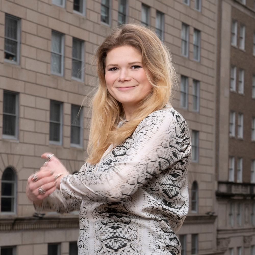 Headshot of a smiling young woman with her arms crossed and standing in front of city backdrop