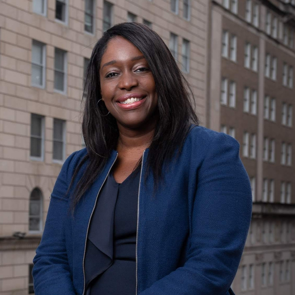 Headshot of a smiling adult woman dressed in business wear sitting in front of a city backdrop