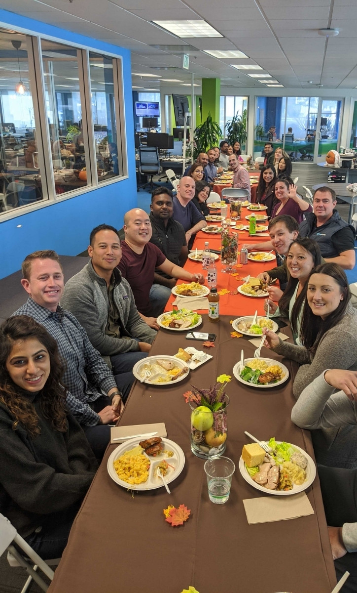 A crowd of smiling PubMatic employees seated at several dinner tables. They all have plats of food in front of them.