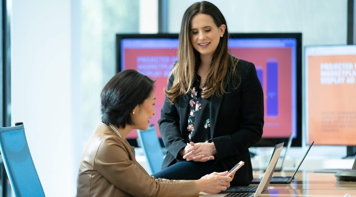 A smiling adult woman sitting on a desk with two large monitors behind her. There's a younger woman sitting in the chair next to her holding a cell phone. She also has a laptop on in front of her