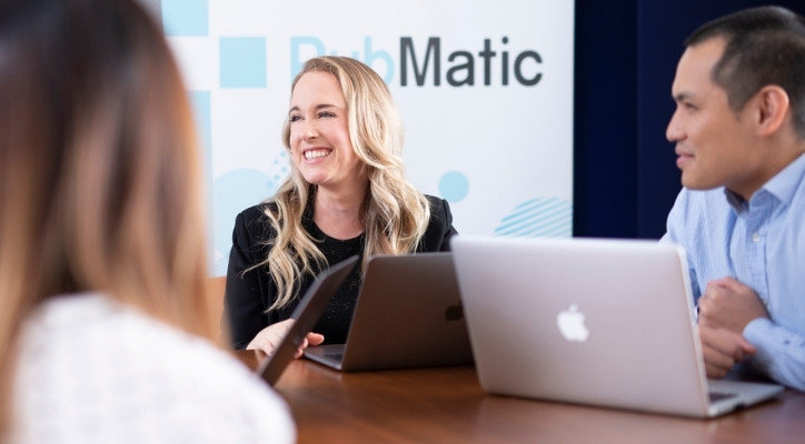 A smiling woman sitting at her desk with her laptop open. There are two other adults next to her with laptops as well