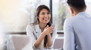 A smiling woman in an office space pointing at the man seated in front of her.