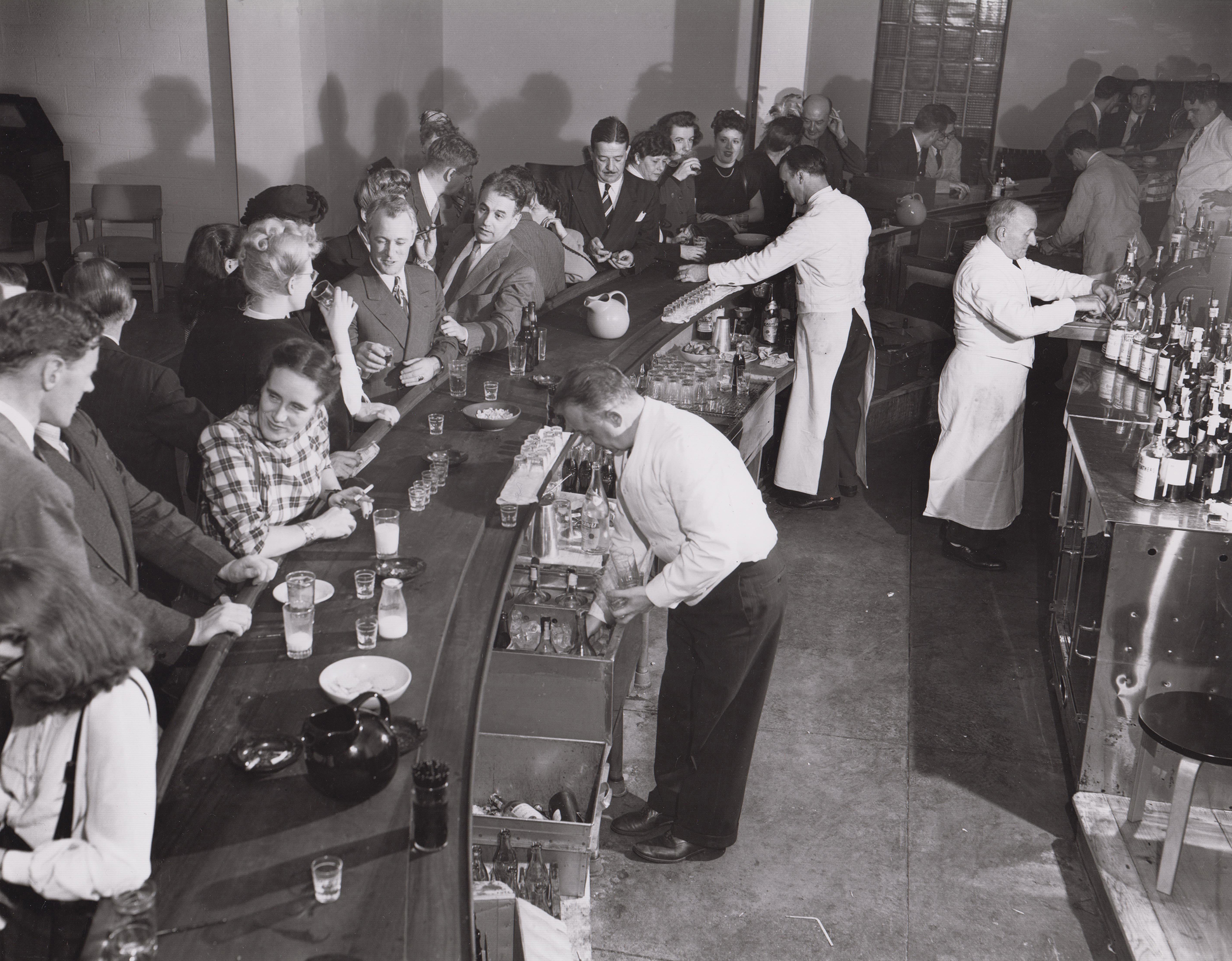 A black and white photo of a rounded bar. On the inside, barkeeps prepare drinks and manage orders. On the outside, patrons stand shoulder to shoulder, engaged in each other’s company. The patrons are fair skinned and a mix of men and women. On the table sits shot glasses, snack bowls, and pitchers. 