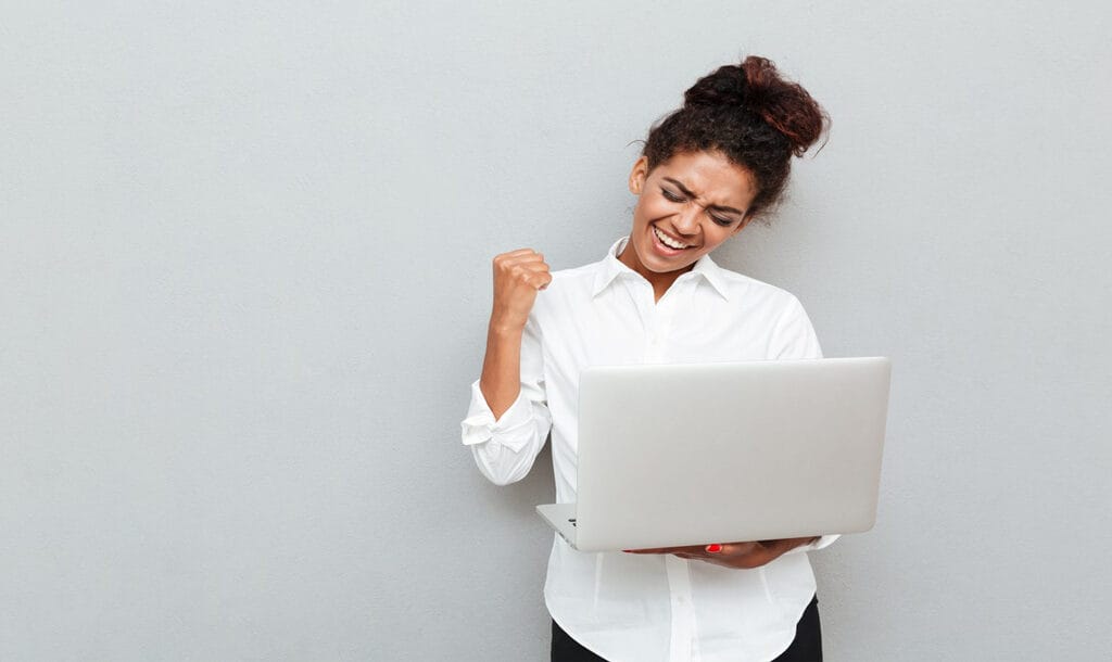 Photo of a cheerful businesswoman standing against a grey wall, holding a laptop, and celebrating a win.