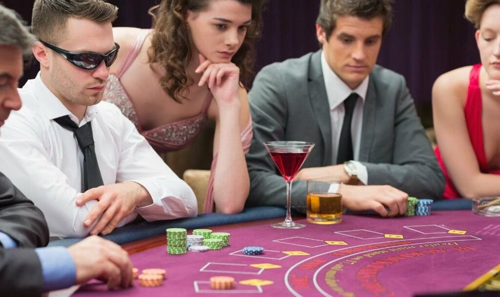 Five well dressed people sitting at a purple felt poker table playing poker waiting for cards to be dealt, two drinks sit on the table