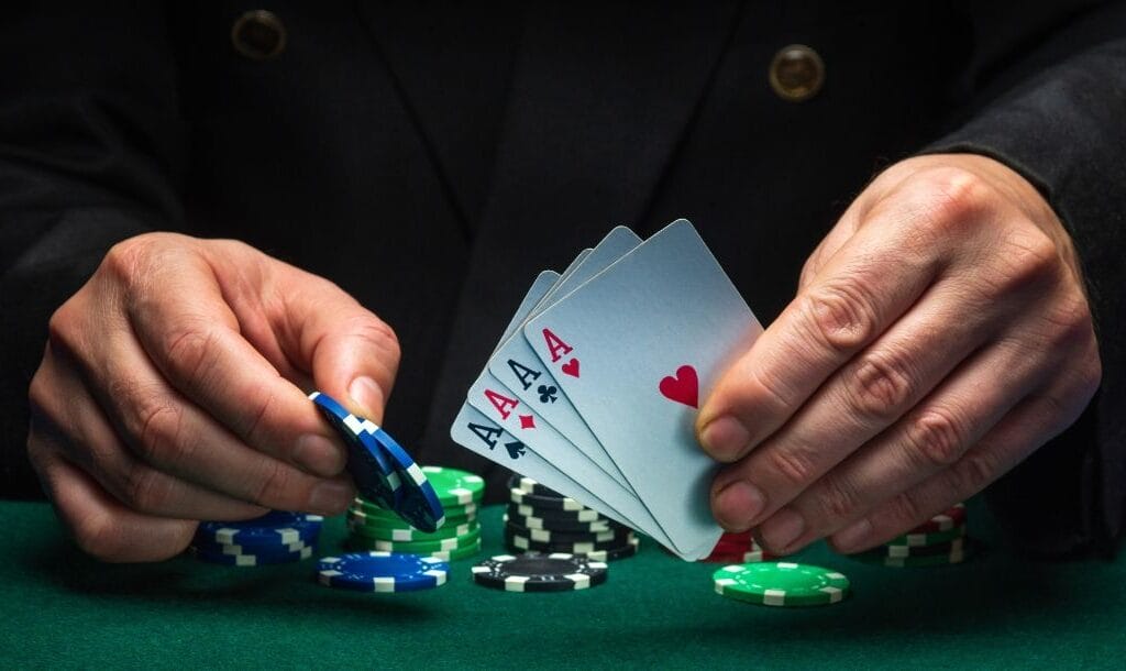 a man holds four of a kind ace playing cards in his left hand and two blue poker chips in his right hand over a green felt poker table with poker chips stacked on it