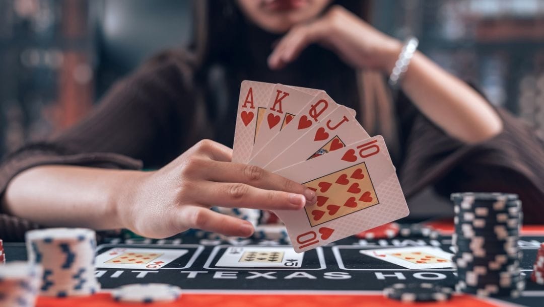 A person sitting at a poker table, revealing a full suit of hearts playing cards, with cards and black poker chips arranged on the poker table.