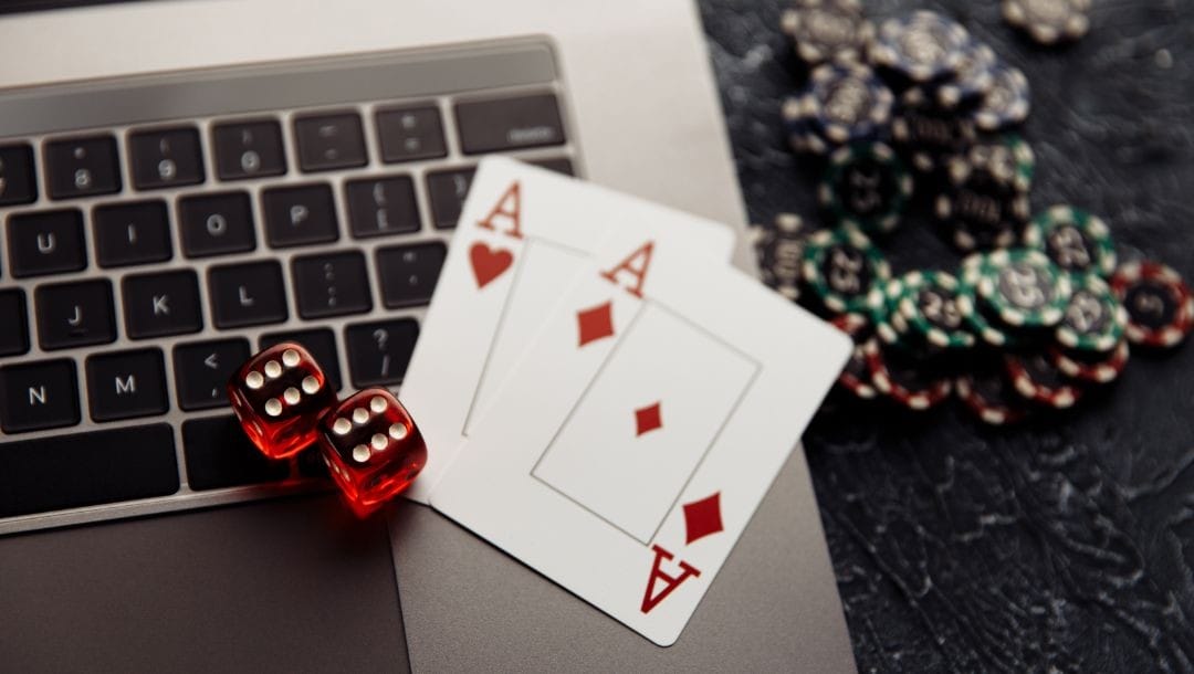 An Ace of Diamonds, an Ace of Hearts, and red dice arranged on a laptop, with casino chips on a table.