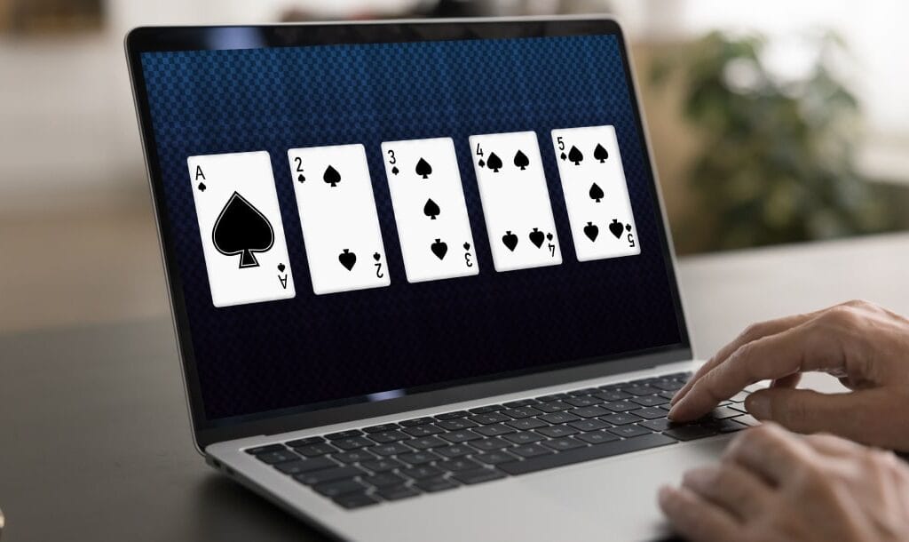 A laptop on a wooden table with five playing cards on the screen. There are hands on the laptop keyboard.