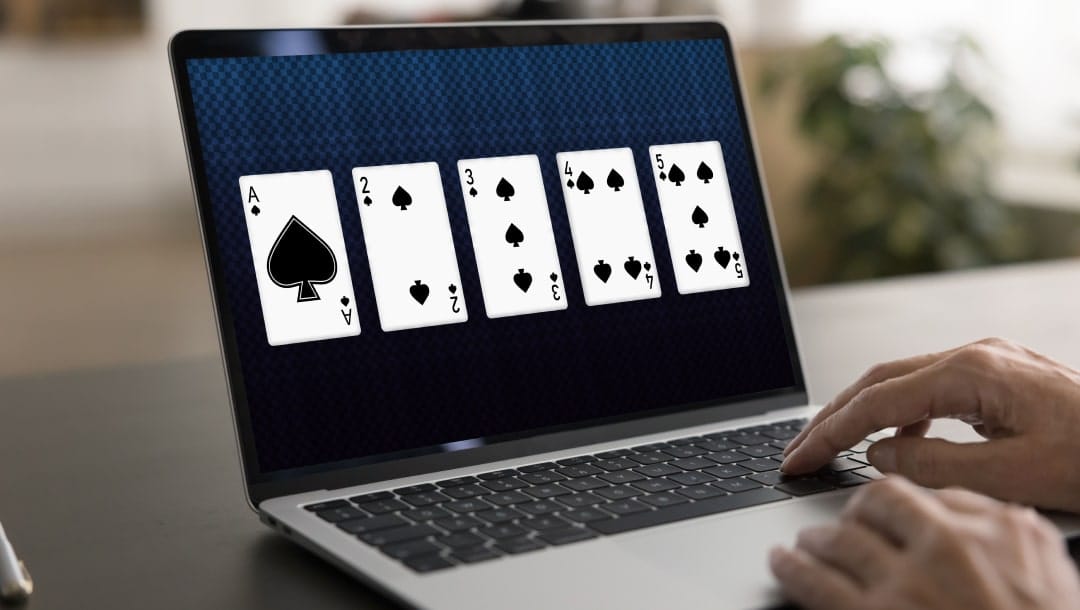 A laptop on a wooden table with five playing cards on the screen. There are hands on the laptop keyboard.