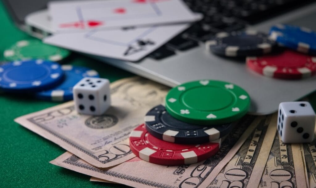 A closeup photograph of $50 bills under the corner of a laptop with poker chips, dice, and playing cards on top of them.