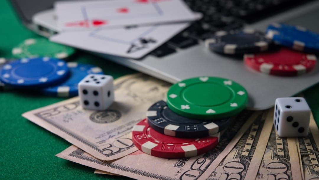 A closeup photograph of $50 bills under the corner of a laptop with poker chips, dice, and playing cards on top of them.