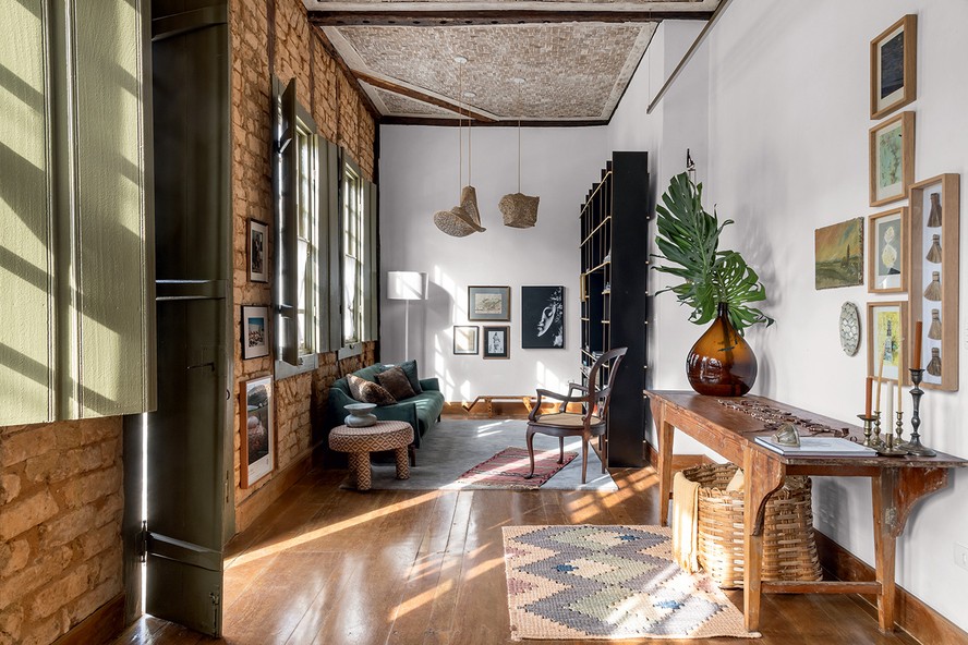 SALA | A estante desenhada pelos arquitetos dá um toque moderno, assim como o sofá garimpado, provavelmente da fábrica L’Atelier, que foi de Jorge Zalszupin, e ganhou companhia dos tapetes sobrepostos da Botteh. Os pendentes de palha de carnaúba são do Projeto Sertões. Sobre o aparador, o vidro âmbar pertenceu à farmácia da família