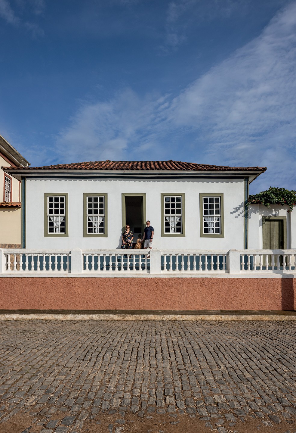 FACHADA | Os proprietários Heloisa Vidigal e Leonardo Lamego estão com a golden retriever Moma em frente ao casarão, cuja fachada original foi mantida. Os batentes das janelas têm uma parte de serralheria com vidro, executada por Zezinho Basílio, e foram pintados em um tom de verde da Suvinil inspirado nos azulejos de Calu Fontes presentes na cozinha — Foto: Evelyn Müller / Divulgação