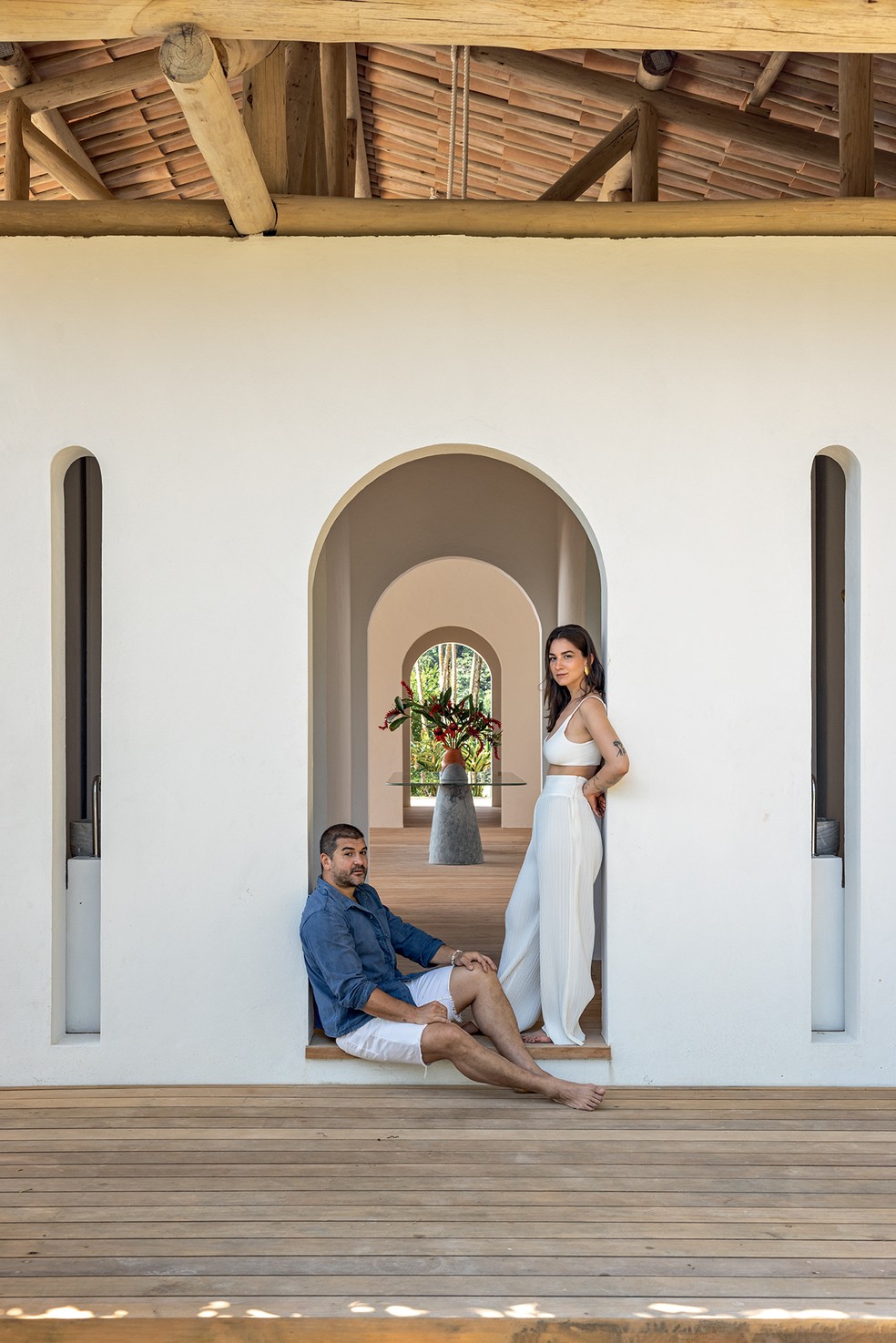 Seba e Luísa, idealizadores do projeto, posam na entrada, em um dos arcos que a atravessam – ao fundo, mesa Mendes, do Estúdio Orth, de pedra-sabão esculpida com tampo de quartzito Botanic Green, da Pettrus — Foto: Fran Parente